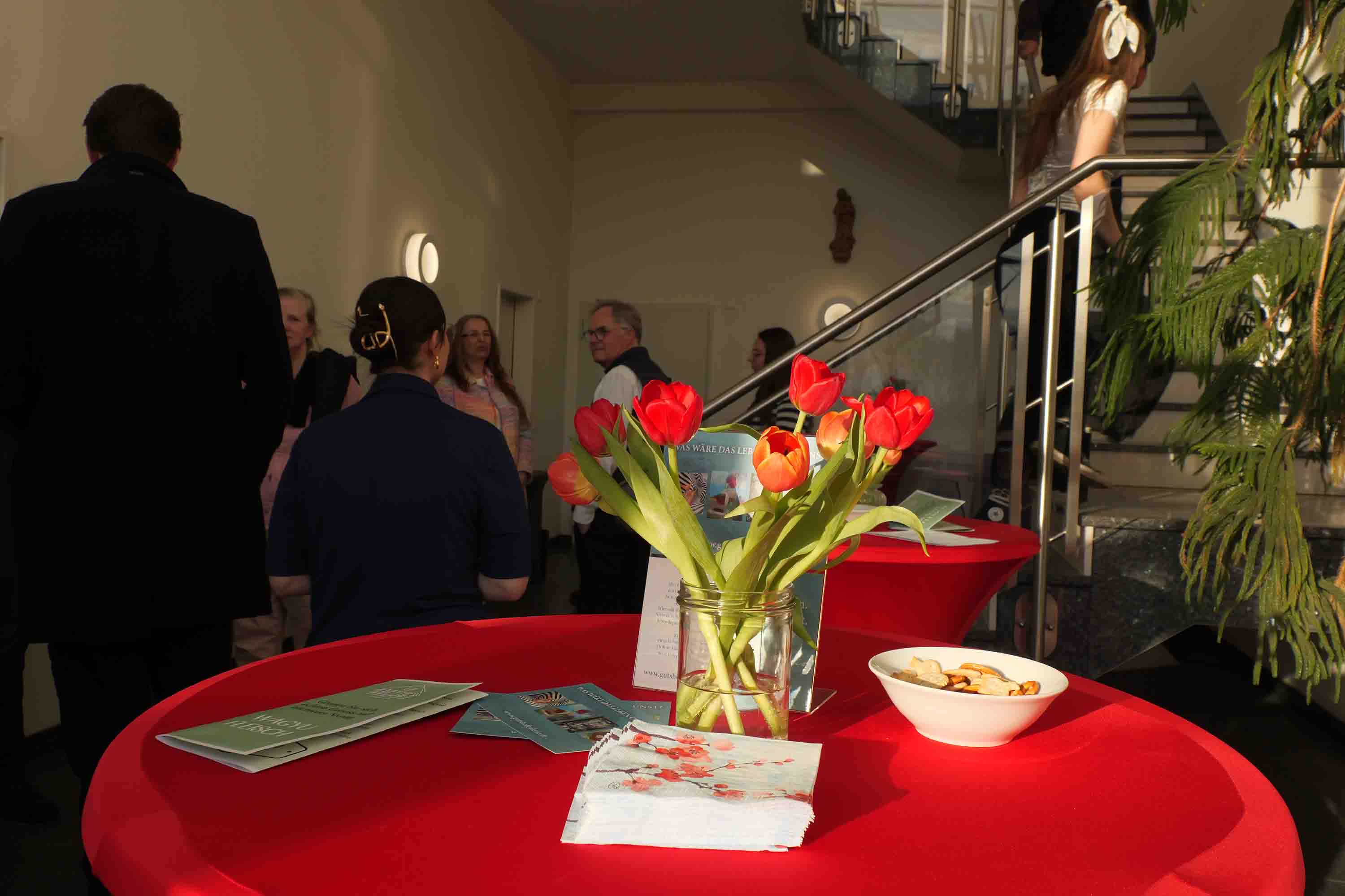 Innenaufnahme einer Veranstaltung: Auf einem runden Tisch mit roter Tischdecke stehen eine Vase mit roten Tulpen, Flyer und eine Schale mit Snacks. Im Hintergrund unterhalten sich mehrere Personen im Foyer eines Geb&auml;udes, w&auml;hrend eine Treppe nach oben f&uuml;hrt.