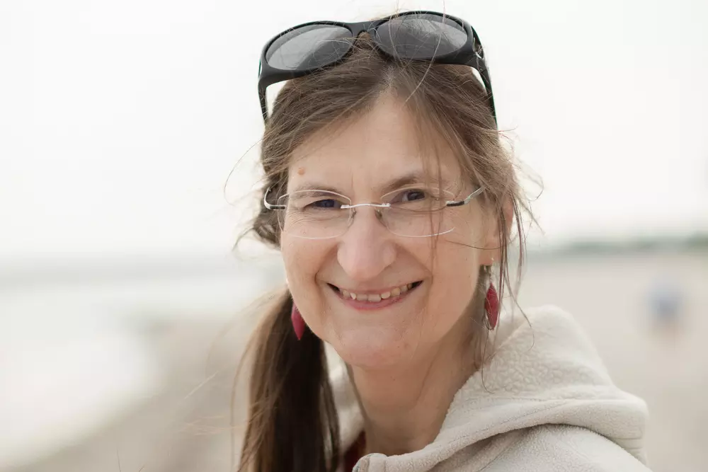 Katrin Mescheder lächelt in die Kamera. Die Frau trägt eine Brille, eine Sonnenbrille sitzt auf ihrem Kopf. Sie steht am Strand, der Hintergrund ist unscharf. Ihre Haare sind leicht vom Wind verweht.